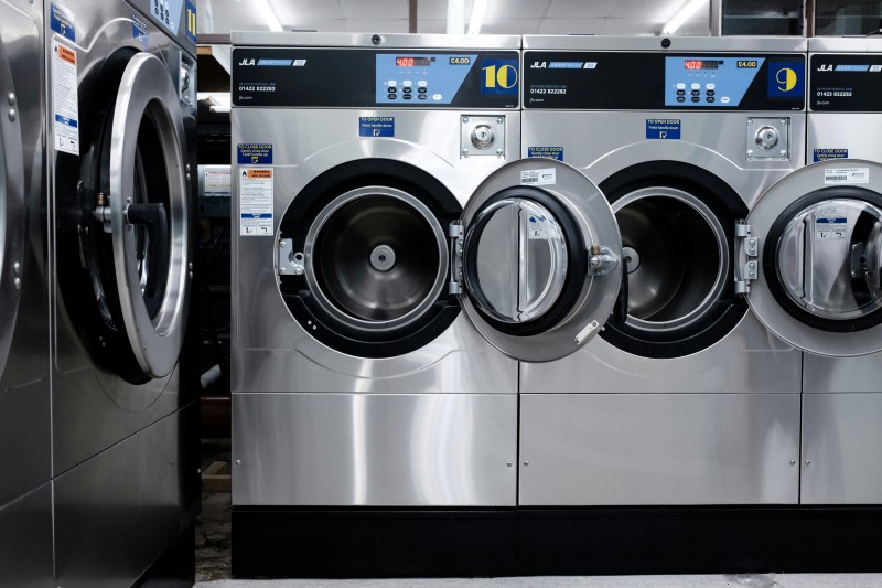 Row of stainless steel commercial washing machines with front-loading doors open in a laundromat.